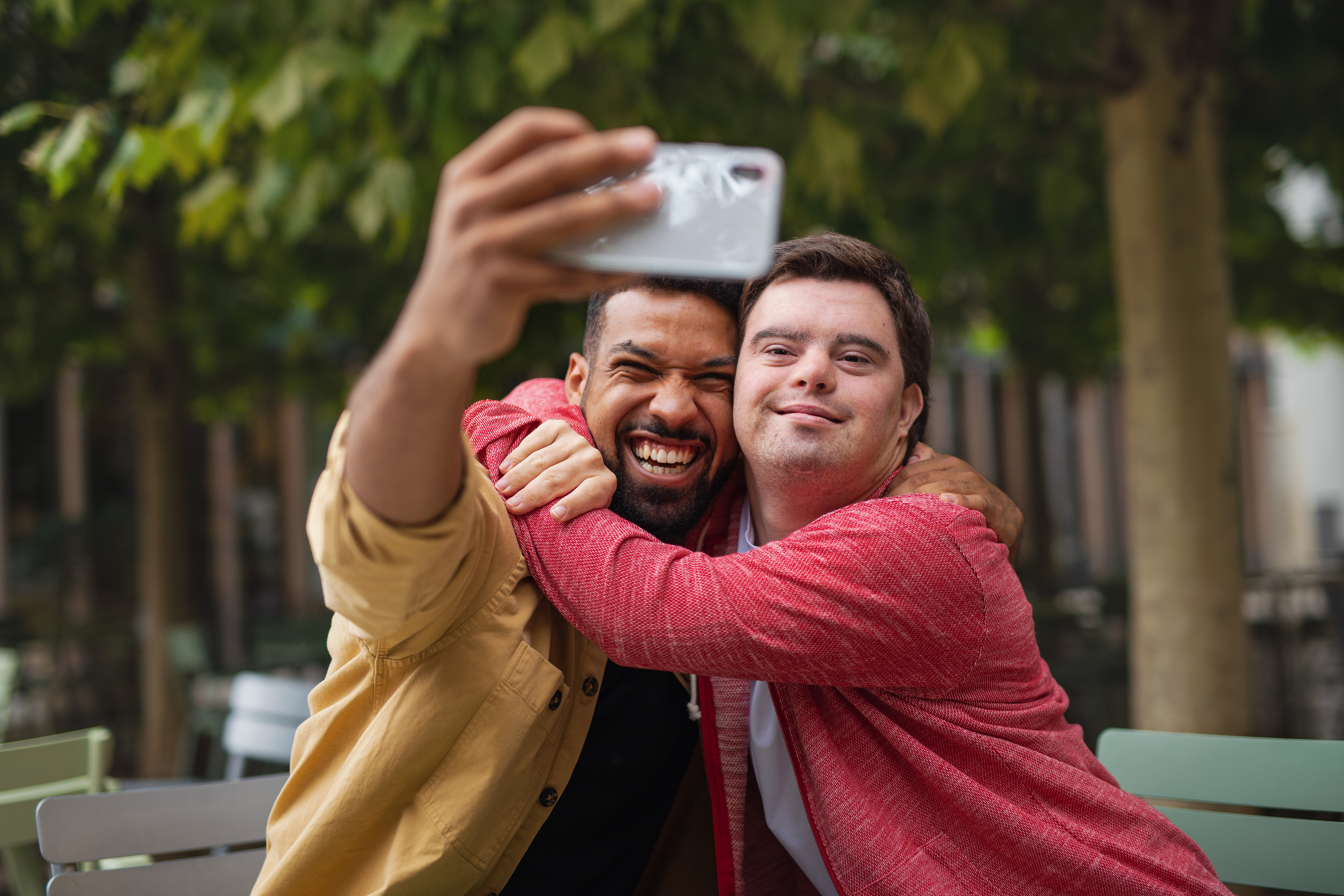 A young man with Down syndrome and his mentoring friend sitting and taking selfie outdoors in cafe