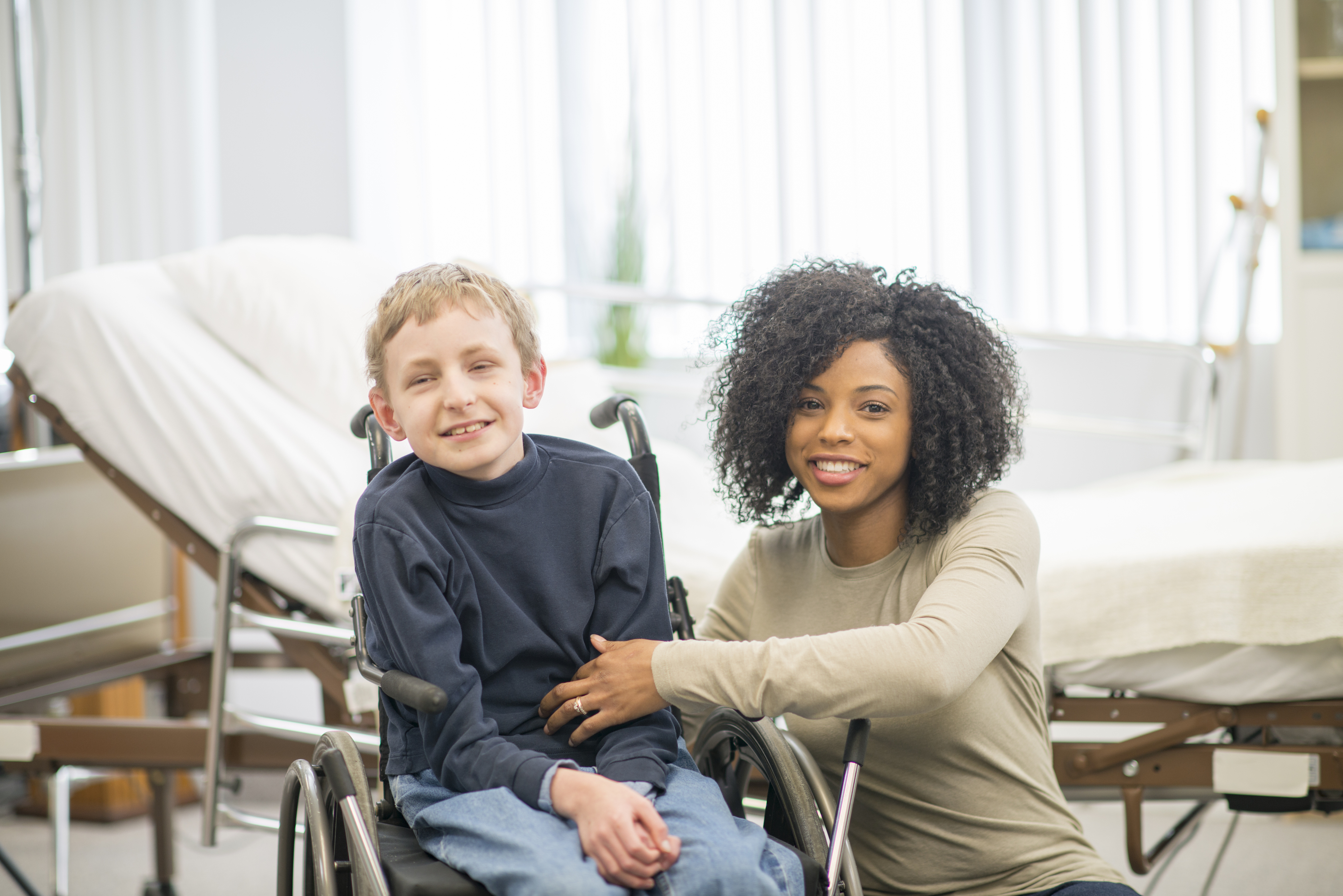 A nurse or caregiver is helping a boy with cerebral palsy get into his wheelchair. They are both smiling and looking at the camera.