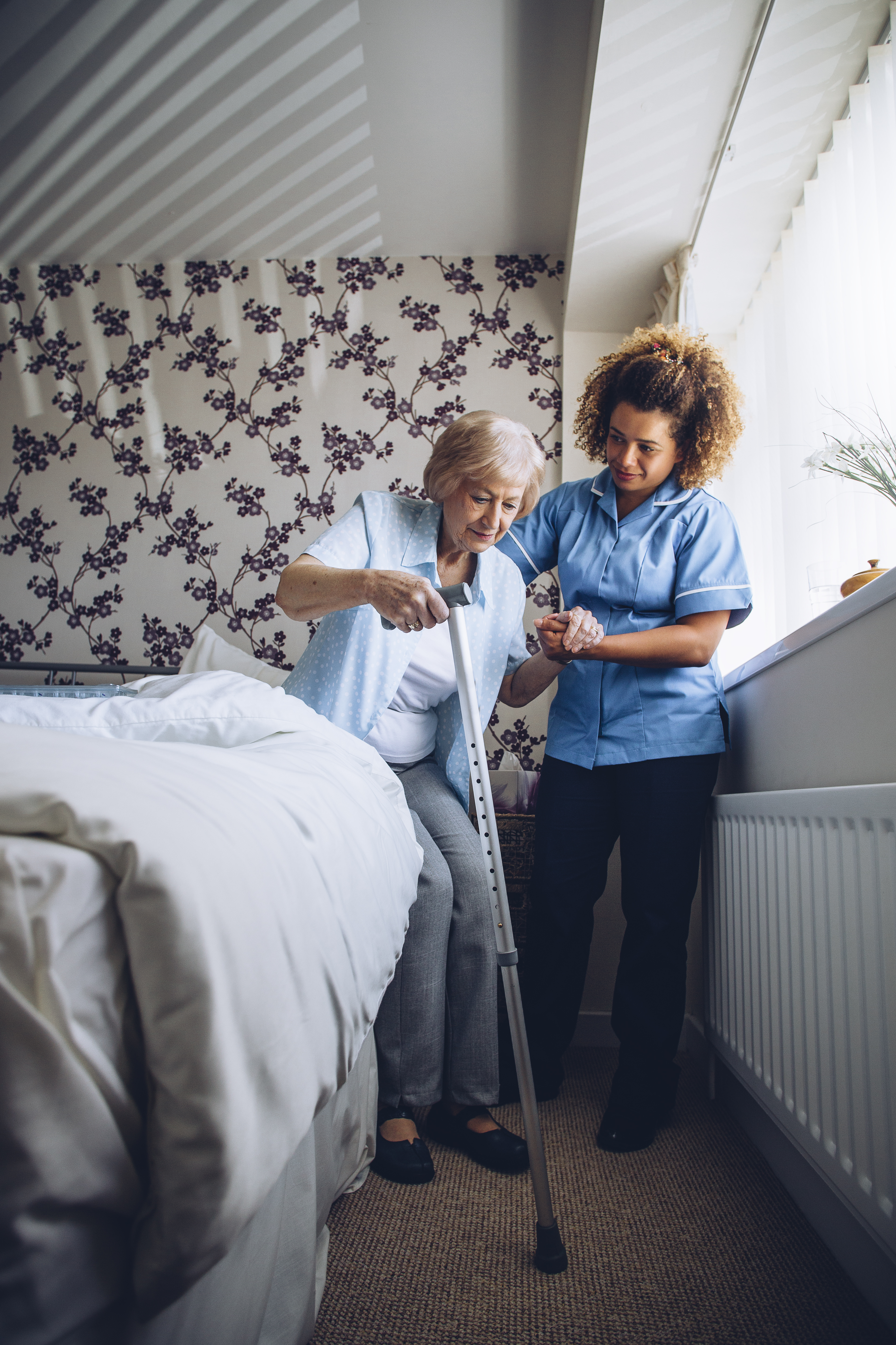 Home Caregiver helping a senior woman get up from her bed with a walking stick.